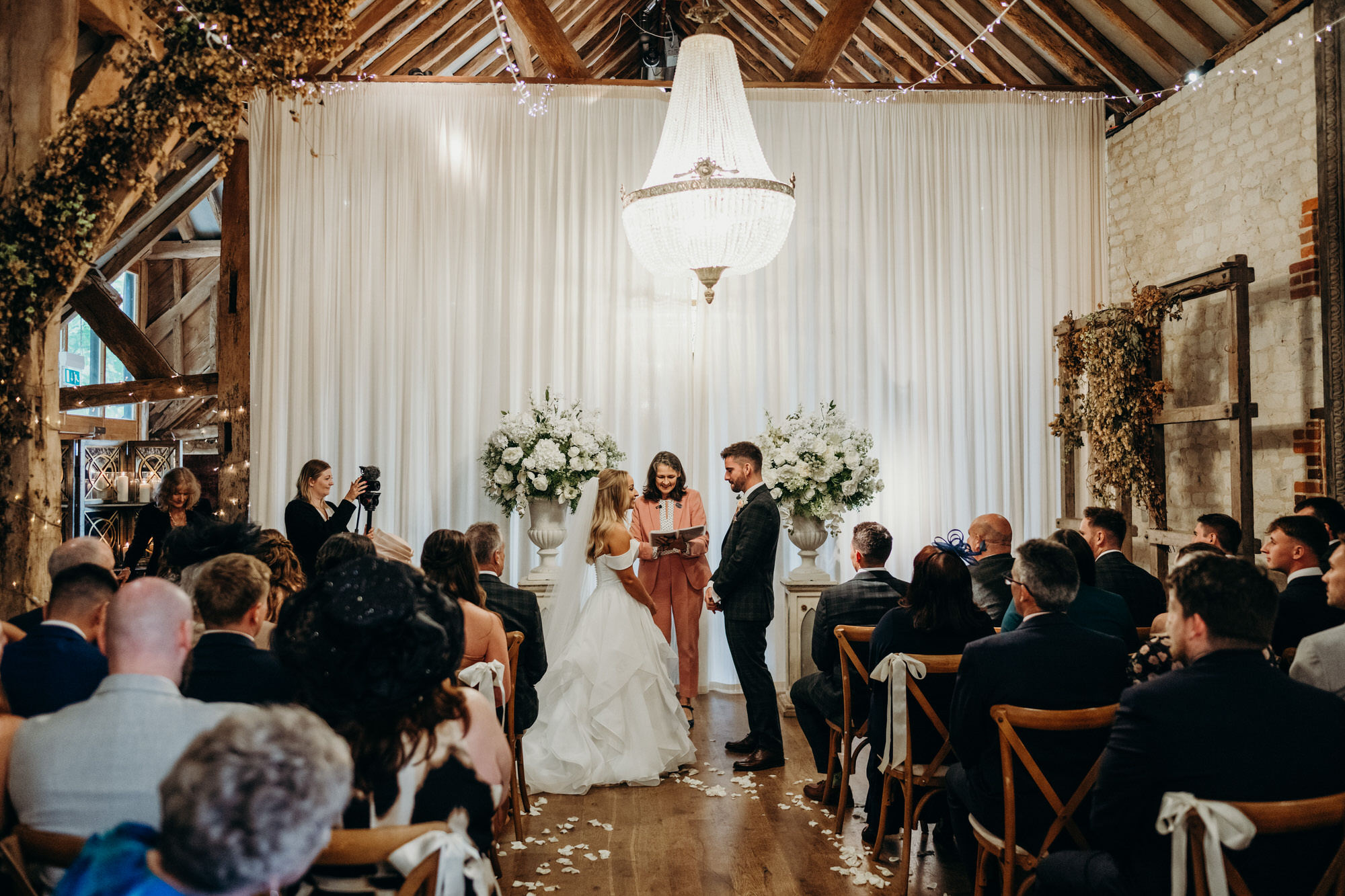 Couple exchanging vows inside Bury Court Barn with warm autumnal floral arrangements and rustic wooden beams.